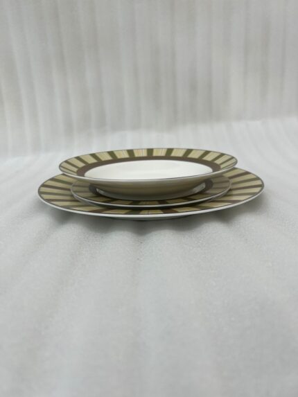 A stacked 18-piece super bone china plate set neatly arranged on a beige linen placemat over a wooden dining table. The ceramic dinnerware set features 6 quarter plates stacked on top, 6 deep soup plates in the middle, and 6 large rice plates at the bottom. Each plate showcases an elegant radiating striped pattern rim in olive green, cream, and taupe tones with fine gold rim detailing. A blur background shows a window and a small flower vase