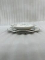 A neatly stacked 18-piece super bone china dinner plate set featuring a delicate grey and blue floral design on a beige table runner alongside a silver spoon and blue flowers.