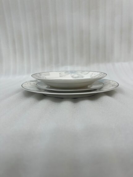 A neatly stacked 18-piece super bone china dinner plate set featuring a delicate grey and blue floral design on a beige table runner alongside a silver spoon and blue flowers.