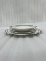 A stacked 18-piece white super bone china plate set with elegant silver-grey floral borders, displayed on a dark wooden table next to a folded cloth napkin with a knife and fork