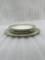 A neatly stacked 18-piece super bone china dinnerware set on a dark wooden dining table. The set includes large rice plates on the bottom, deep plates in the middle, and smaller quarter plates on top, all featuring an elegant light green and gold brushstroke pattern around the borders. Green linen napkins and silver cutlery are arranged neatly beside the stack.