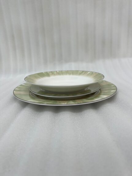 A neatly stacked 18-piece super bone china dinnerware set on a dark wooden dining table. The set includes large rice plates on the bottom, deep plates in the middle, and smaller quarter plates on top, all featuring an elegant light green and gold brushstroke pattern around the borders. Green linen napkins and silver cutlery are arranged neatly beside the stack.
