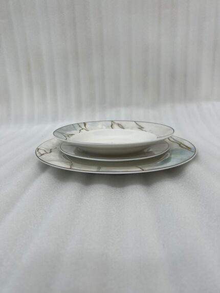 A neatly stacked 18-piece super bone china dinner plate set featuring a white and mint green marble pattern with gold veining, arranged on a dining table with silverware and a crystal glass.