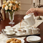 A high-angle shot of a hand pouring hot tea from a white gold-rimmed bone china teapot into a matching cup on a silver tray, surrounded by pastries and a floral centerpiece.