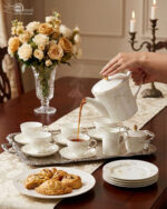 A high-angle shot of a hand pouring hot tea from a white gold-rimmed bone china teapot into a matching cup on a silver tray, surrounded by pastries and a floral centerpiece.