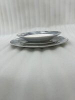 A neatly stacked 18-piece luxury super bone china plate set on a dining table runner. The dinnerware features a contemporary blue and gray ocean marble pattern with subtle gold vein accents and a thin silver rim. The stack consists of three sizes: small quarter plates on top, medium deep soup plates in the middle, and large flat rice plates at the bottom, displayed in a brightly lit, elegant dining room setting with candlesticks in the background.