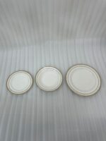 A stacked 18-piece white Super Bone China dinner plate set with elegant gold borders, arranged neatly on a polished wooden dining table alongside a wine glass and silverware under warm chandelier lighting.