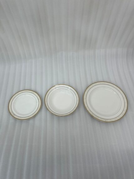 A stacked 18-piece white Super Bone China dinner plate set with elegant gold borders, arranged neatly on a polished wooden dining table alongside a wine glass and silverware under warm chandelier lighting.