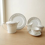 A complete 24-piece luxury tea set on a wooden table, featuring a white porcelain teapot, milk jug, sugar pot, cups, saucers, and plates with delicate silver fern patterns.
