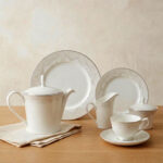 A complete 24-piece white porcelain tea set featuring delicate lace-patterned edges and silver trim, arranged on a wooden table.
