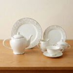 A 24-piece white porcelain tea set featuring a delicate silver floral scroll pattern, including a teapot, cups, saucers, plates, and serving pots, arranged on a wooden table.