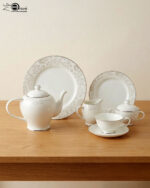 A 24-piece white porcelain tea set featuring a delicate silver floral scroll pattern, including a teapot, cups, saucers, plates, and serving pots, arranged on a wooden table.