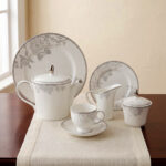 A sophisticated 24-piece white porcelain tea set featuring a delicate grey floral pattern and silver trim, arranged elegantly on a table runner.