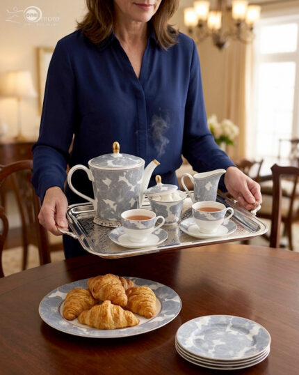 A woman in a navy blue blouse holding a silver tray with a floral blue and white Super Bone China tea set, including a teapot, cups, and sugar bowl, with a plate of croissants in the foreground