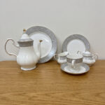 : A complete 24-piece white porcelain tea set featuring delicate grey floral patterns with gold trim, including a teapot, teacups, saucers, and plates, arranged on a wooden surface.