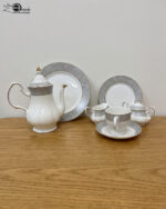 : A complete 24-piece white porcelain tea set featuring delicate grey floral patterns with gold trim, including a teapot, teacups, saucers, and plates, arranged on a wooden surface.
