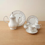 A 24-piece white porcelain tea set featuring delicate gray floral patterns and silver trim, arranged on a light wooden surface.