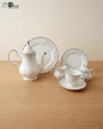 A 24-piece white porcelain tea set featuring delicate gray floral patterns and silver trim, arranged on a light wooden surface.