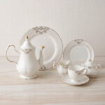 A luxurious 24-piece white porcelain tea set featuring delicate gold filigree patterns, including a teapot, teacups, saucers, plates, and serving pieces displayed on a marble-textured surface
