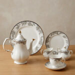 A 24-piece luxury porcelain tea set featuring a white teapot, milk jug, sugar pot, cups, saucers, and plates adorned with delicate gray floral patterns and gold rims on a wooden table.