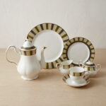 : A complete 24-piece porcelain tea and dining set featuring white ceramic with gold and olive-green striped rims, including plates, cups, saucers, a teapot, a sugar bowl, and a milk creamer arranged on a wooden surface.