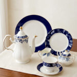 A 24-piece luxury porcelain tea set featuring a classic blue and white floral pattern with gold trim, including a teapot, cups, saucers, and plates, arranged on a beige table runner.