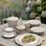 A premium Super Bone China tea set featuring a teapot, sugar bowl, creamer, and cups and saucers with a tan pinstripe pattern and gold-rimmed borders, arranged on a wooden table outdoors with avocado toast on a plate.