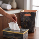 A close-up photograph of a hand pulling a tissue from a luxury black marble and gold-veined tissue box cover with gold trim, next to a matching small open-top waste bin with a warm terracotta interior and cutout handles, all on a wooden surface in a contemporary bathroom setting.
