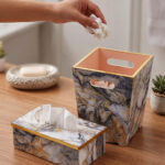 A hand dropping a tissue into a modern marble-patterned dustbin with gold trim, sitting next to a matching tissue box cover on a wooden counter.