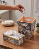 A hand dropping a tissue into a modern marble-patterned dustbin with gold trim, sitting next to a matching tissue box cover on a wooden counter.