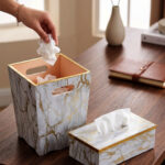 A close-up view of a luxury faux-marble waste bin with intricate gold veining and polished gold trim, positioned next to a matching marble tissue box holder on a wooden desk. A hand is seen depositing a tissue into the bin, which features a peach-pink interior lining.