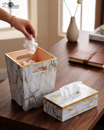 A close-up view of a luxury faux-marble waste bin with intricate gold veining and polished gold trim, positioned next to a matching marble tissue box holder on a wooden desk. A hand is seen depositing a tissue into the bin, which features a peach-pink interior lining.