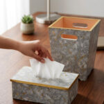 A close-up view of a hand reaching to pull a tissue from a grey and gold-veined faux-marble tissue box cover with polished gold trim, next to a matching compact grey marble waste bin with handle cutouts and a peach-colored interior, sitting on a wooden desk.