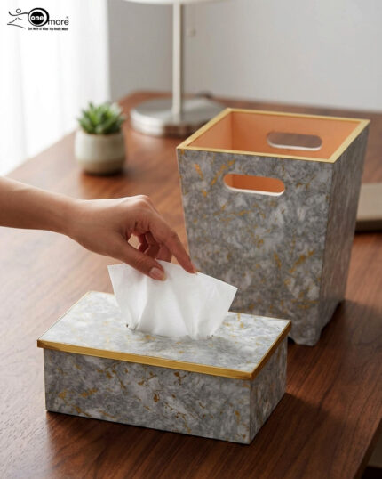 A close-up view of a hand reaching to pull a tissue from a grey and gold-veined faux-marble tissue box cover with polished gold trim, next to a matching compact grey marble waste bin with handle cutouts and a peach-colored interior, sitting on a wooden desk.