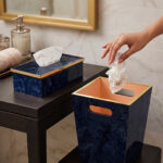 A luxury blue marble resin tissue box cover and matching wastebasket with elegant gold trim on dark wooden tables in a bathroom setting.