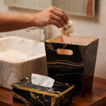 Luxury black and gold marble pattern dustbin and matching tissue box holder on a wooden vanity next to a marble bathroom sink.