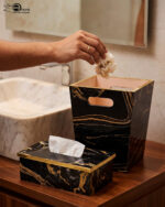 Luxury black and gold marble pattern dustbin and matching tissue box holder on a wooden vanity next to a marble bathroom sink.
