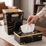 A hand pulling a tissue from a luxury black and gold marble tissue box cover next to a matching wastebasket on a table.