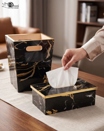 A hand pulling a tissue from a luxury black and gold marble tissue box cover next to a matching wastebasket on a table.