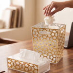 Clear acrylic tissue box holder and matching small desktop dustbin with an elegant gold geometric lattice pattern on a wooden desk.