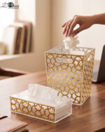 Clear acrylic tissue box holder and matching small desktop dustbin with an elegant gold geometric lattice pattern on a wooden desk.