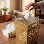An elegant gold and clear acrylic dust bin with a laser-cut leaf pattern on a wooden table, featuring a matching tissue box holder and a hand dropping a crumpled tissue inside.