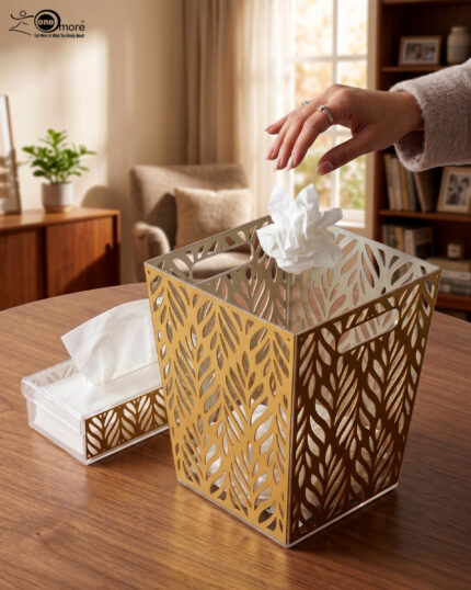 An elegant gold and clear acrylic dust bin with a laser-cut leaf pattern on a wooden table, featuring a matching tissue box holder and a hand dropping a crumpled tissue inside.