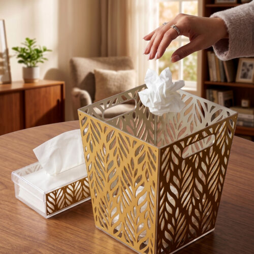 An elegant gold and clear acrylic dust bin with a laser-cut leaf pattern on a wooden table, featuring a matching tissue box holder and a hand dropping a crumpled tissue inside.
