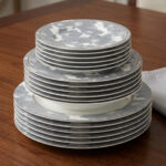 A stacked 18-piece super bone china dinnerware set featuring a grey and white floral pattern on a wooden table, including rice, deep, and quarter plates.