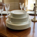 A stacked 18-piece white super bone china plate set on a wooden dining table, featuring 6 rice plates, 6 deep plates, and 6 quarter plates with a subtle design