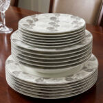 Stacked 18-piece super bone china plate set with grey floral patterns, including rice plates, deep plates, and quarter plates on a wooden dining table.