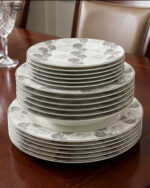 Stacked 18-piece super bone china plate set with grey floral patterns, including rice plates, deep plates, and quarter plates on a wooden dining table.