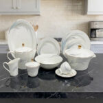 An elegant 61-piece white Super Bone China dinner set with blue feather patterns and gold accents, displayed on a black marble countertop in a kitchen