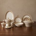 A premium 61-piece Super Bone dinner set by OneMore Crockery featuring cream-colored plates with gold-rimmed textured borders, including a teapot, cups, serving bowls, and platters arranged on a dark wooden table
